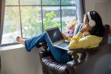 Wall Mural - Businesswoman working online at the home office via laptop. Asian young entrepreneur watching webinars and talking during meeting video conferences call with team. Woman sitting at sofa in living room