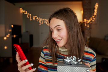 Happy adult female with gift box smiling and browsing social media on smartphone while celebrating birthday at home