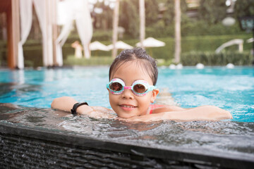 Wall Mural - smiling child wearing swimming glasses in swimming pool. little girl playing in outdoor swimming pool on summer vacation on tropical beach island. child learning to swim in pool of luxury resort.