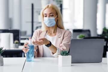 Wall Mural - Personal hygiene in modern office after lockdown. Attractive woman in protective mask and smart watch disinfect hands with antiseptic at work with laptop