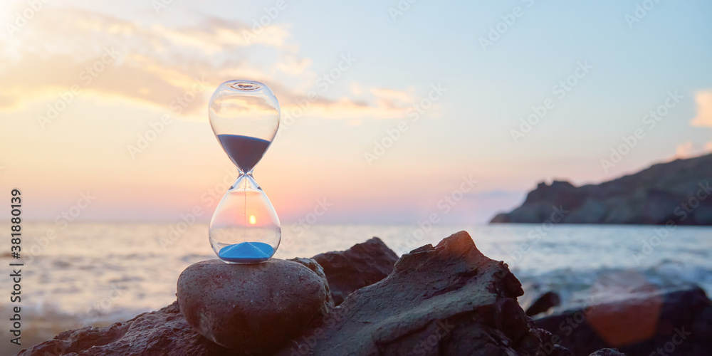 Hourglass with blue falling sand inside. Ocean landscape and golden hour. Time fly and need in rest concept, copy space - obrazy, fototapety, plakaty 