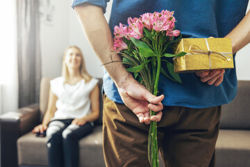 Wall Mural - husband hiding romantic surprise present and flowers behind back to beloved wife at home