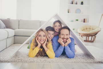 Wall Mural - Safe family new house concept. Photo of people mom dad two small kids lying floor under paper card roof in living room indoors