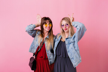 Lifestyle studio image of two cheerful cute women,  best friends having great time together , posing over pink background . Wearing jeans jackets and elegant dress .