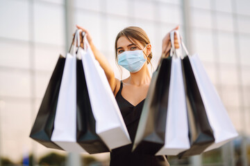 Young woman in protective sterile medical mask on her face with shopping bags near shopping center. Shopping during the coronavirus Covid-19 pandemic. Purchases, black friday, discounts, sale concept.