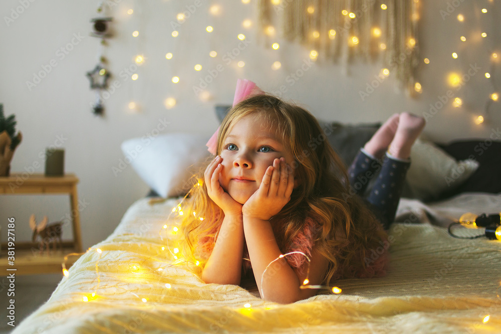A happy little girl with blonde hair and blue eyes is lying on the bed ...