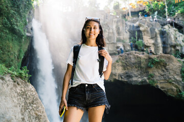 Wall Mural - Cheerful female traveler with phone standing against waterfall