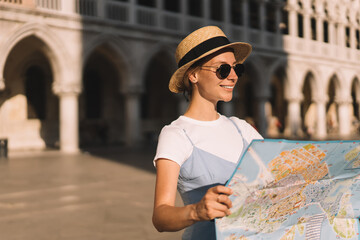 Wall Mural - Happy smiling tourist in stylish sunglasses and straw hat using paper navigation map for search showplaces in romantic Italian city - Venezia, Caucasian woman choosing showplace for visiting