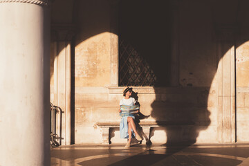 Wall Mural - Cheerful woman holding map and sitting in sunny arched gallery