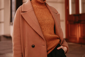 young woman with cropped head in the orange knitted cozy sweater and brown coat standing on the stre