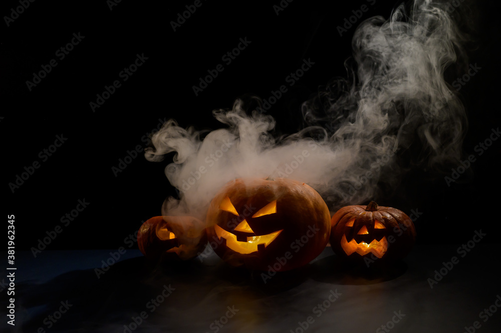 Three creepy halloween steaming pumpkins with a carved luminous smirk on a black background. A handmade jack-o-lantern head with a candle inside in the dark among the fog. Trick or treat. Postcard. - obrazy, fototapety, plakaty 