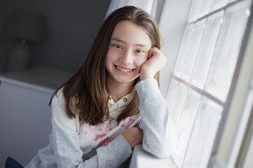 young teenage girl sitting at the window smiling with her hand against her face