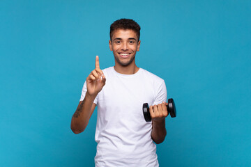 Wall Mural - young man lifting a sumbbell smiling and looking friendly, showing number one or first with hand forward, counting down