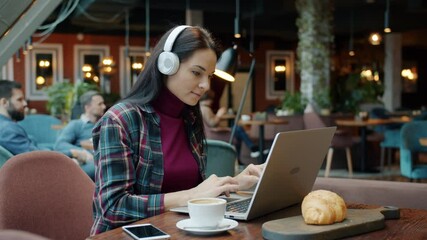 Female freelancer is working with laptop computer typing and enjoying music with headphones in cafe. Modern lifestyle and technology concept.