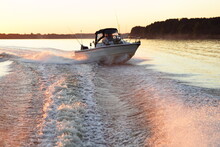 Fast Gliding Motor Boat In Water Foam Track On Beautiful Sunset Background, Bright Orange Sunshine In Calm Water On Forest On Horizon At Sunny Summer Evening Bow Side Front View From Powerboat Transom