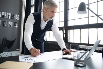 Pensive handsome matured male owner of corporation reading financial documentation standing near table, confident businessman in trendy formal wear checking accounting report