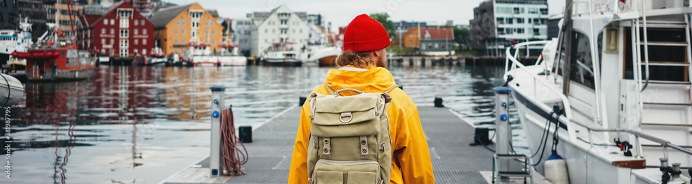 Man tourist with touristic rucksack wearing yellow jacket walking among authentic fishing boats. Wide image - obrazy, fototapety, plakaty 
