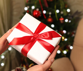 Women's hands hold a white gift box with a red ribbon on the background of a Christmas tree. The concept of New year and a happy Christmas