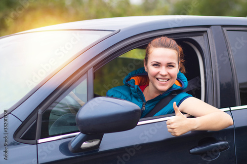 Happy woman, driver, young positive man, driving her car, smiling shows thumb up as a gesture from the car window. Happy buyer, new car buyer enjoying driving. Successful purchase of a female