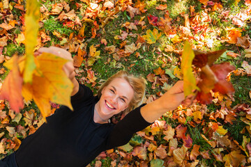 A young woman lies on fallen orange leaves and holds yellow maple leaves in her hand.