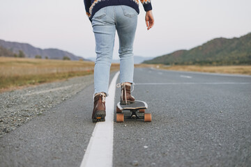 Wall Mural - Crop photo of women's legs in jeans on longboard on road with the mountains background