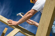 © sculpies - roofer carpenter builder holding a hammer nail working on roof structure construction site outdoors blue sky and yellow hardhat