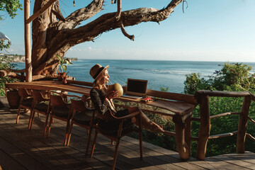 Pretty young woman using laptop in cafe with sea view. Freelancer working remotely from beach cafe, drink coconut and use laptop