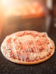 Uncooked peperoni and salami pizza in plastic wrap in home kitchen environment on dark table surface. Selective focus. Sun flare