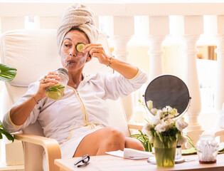 Beautiful woman wearing white bathrobe with cucumber skin care mask while drinking detox drink. DIY home spa