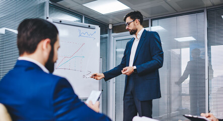 Group of Caucasian colleagues analyzing revenue statistics during co brainstorming, confident formally dressed leaders discussing infographics from flipchart during collaborative working process