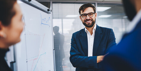 Cheerful male leader of company smiling near flip chart during presentation with colleagues, happy businessman in formal outfit and spectacles discussing economic graphics with cropped partners