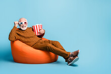 Portrait Of His He Nice Attractive Funky Cheerful Cheery Glad Bearded Grey-haired Man Sitting In Bag Chair Eating Corn Watching Film Having Fun Isolated Over Blue Pastel Color Background