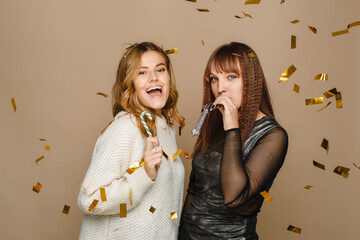 Wall Mural - Two young women on a beige background in a sweater and a black dress are blowing a festive tune and holding a candy cane. Celebrating Christmas and New Year