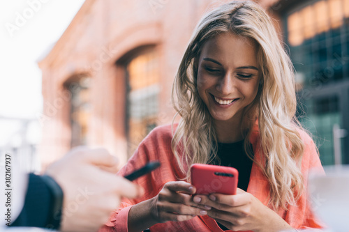 Cheerful caucasian blonde woman holding mobile phone sending and texting messages on leisure,20s cropped image of female sitting on cafe terrasse share information and content via smartphone