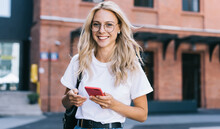 Half Length Portrait Of Cheerful Caucasian Female In Trendy Wear Spending Time On Street Using Smartphone, Beautiful Millennial Hipster Girl Blogger Looking At Camera In Town Holding Mobile Phone