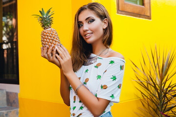 portrait of attractive woman in pinted t-shirt on summer vacation holding pineapple, fruit diet detox, tanned skin, bright yellow background