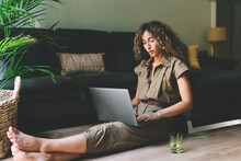 Beautiful Female In Casual Outfit Chilling On A Floor Beside Comfortable Couch In A Living Room With Laptop On Her Knees. Web Designer Browsing  Laptop While Working On A Remote Project At Home.