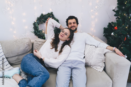 Smiling caucasian male and female couple in love resting on sofa in festive decorated apartment during winter vacations, positive marriage looking at camera celebrating new year evening together