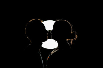 Couple of man and woman looking closely at each other and kissing in the dark illuminated by a light behind them.