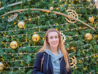 Wall Mural - Smiling girl on the background of a large Christmas tree decorated with a garland and golden balls