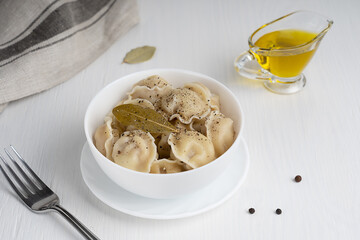 Wall Mural - Pelmeni or dumplings of Russian cuisine made of minced meat filling wrapped in dough served in white bowl with fork with black pepper seasoning and bay leaf on white wooden background. Horizontal