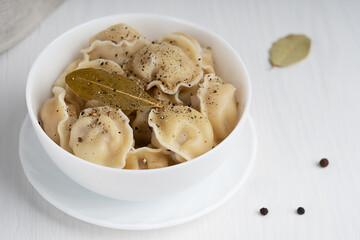 Poster - Pelmeni or dumplings of Russian cuisine made of minced meat filling wrapped in dough served in white bowl with black pepper seasoning and bay or laurel leaf on white wooden background. Horizontal