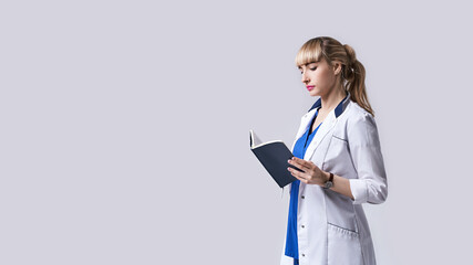 Confident young woman doctor standing with notebook or medical book in her hands. Cheks records, recipes or prescriptions. Intern in white medical clothes posing on light grey background.