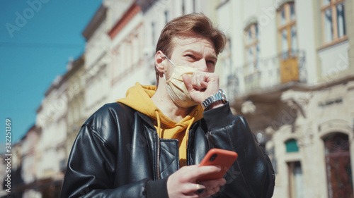 Fotografía Blonde stylish caucasian guy wearing medical mask using smartphone coughing badly covering face suffering symptoms of coronavirus outdoors