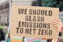The Phrase " We Should Slash Emissions To Net Zero " On A Banner In Men's Hand With Blurred Background. Ecology. Industry. Technology. Pollution. CO2. Global Warming. Decrease
