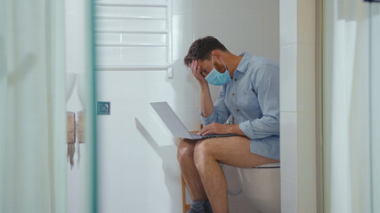 Unhappy worried businessman working on laptop wearing mask sit on the toilet at home restroom bearded wc isolation quarantine pandemic coronavirus covid-19 portrait close up slow motion