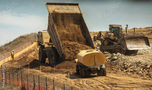 A very large haul dump truck at a construction site dumping a massive amount of dirt near a water truck and a bulldozer