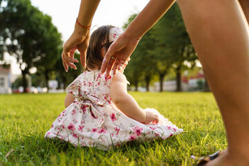 Back view One small caucasian baby girl sitting on the grass wearing dress in the field in summer or autumn day while hands of unknown woman try to reach her - childhood and motherhood concept