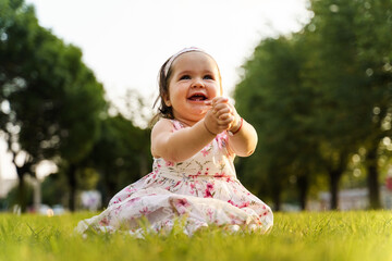 One small caucasian baby girl sitting on the grass wearing dress in the field in summer or autumn day smiling happy having fun clapping hands - childhood concept