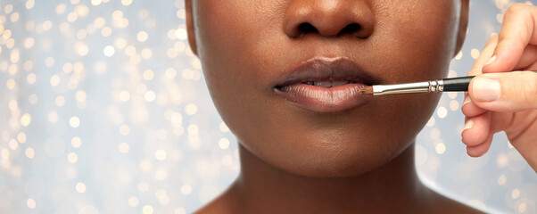 beauty, cosmetics and people concept - close up of face of beautiful young african american woman and hand with make up brush applying lipstick or gloss to lips over festive lights background